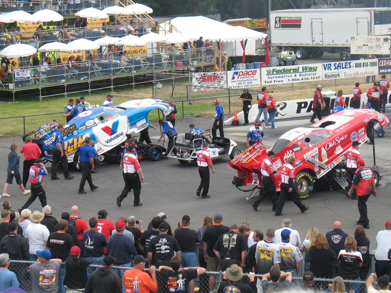 Wide view at Pacific Raceways: Beckman's blue Valvoline Charger Funny Car on the left and a red Funny Car on the right, both with bodies raised and crews around them, spectators lined up along a fence in the foreground